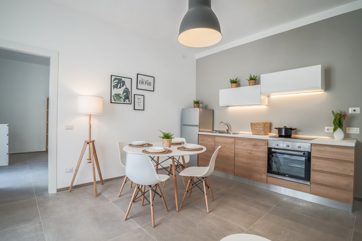 A modern kitchen space is presented featuring light wood cabinetry and sleek appliances. A circular dining table with four white chairs sits in the center, complemented by potted plants on the countertop. Warm lighting enhances the airy ambiance, and a light fixture hangs above the table.