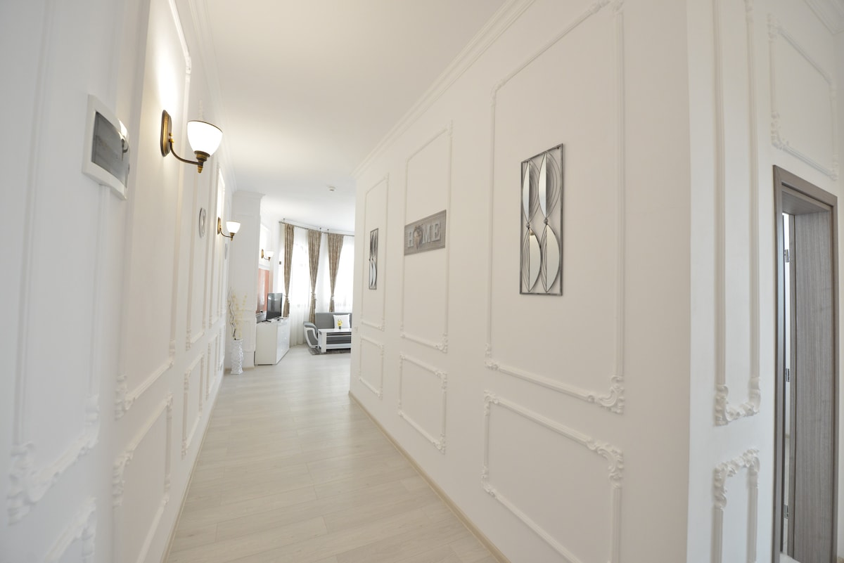 A bright hallway features elegant white walls adorned with framed artwork. Decorative lighting fixtures enhance the space, leading unobtrusively to the living area visible at the end. Light wood flooring complements the modern design, contributing to a spacious feeling.