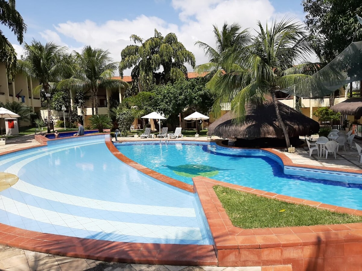 A spacious outdoor pool is surrounded by tropical palm trees and sun loungers. Umbrellas offer shaded seating areas, while thatched-roof structures are visible nearby. The pool's blue tiles reflect the sky, creating a serene environment ideal for relaxation.
