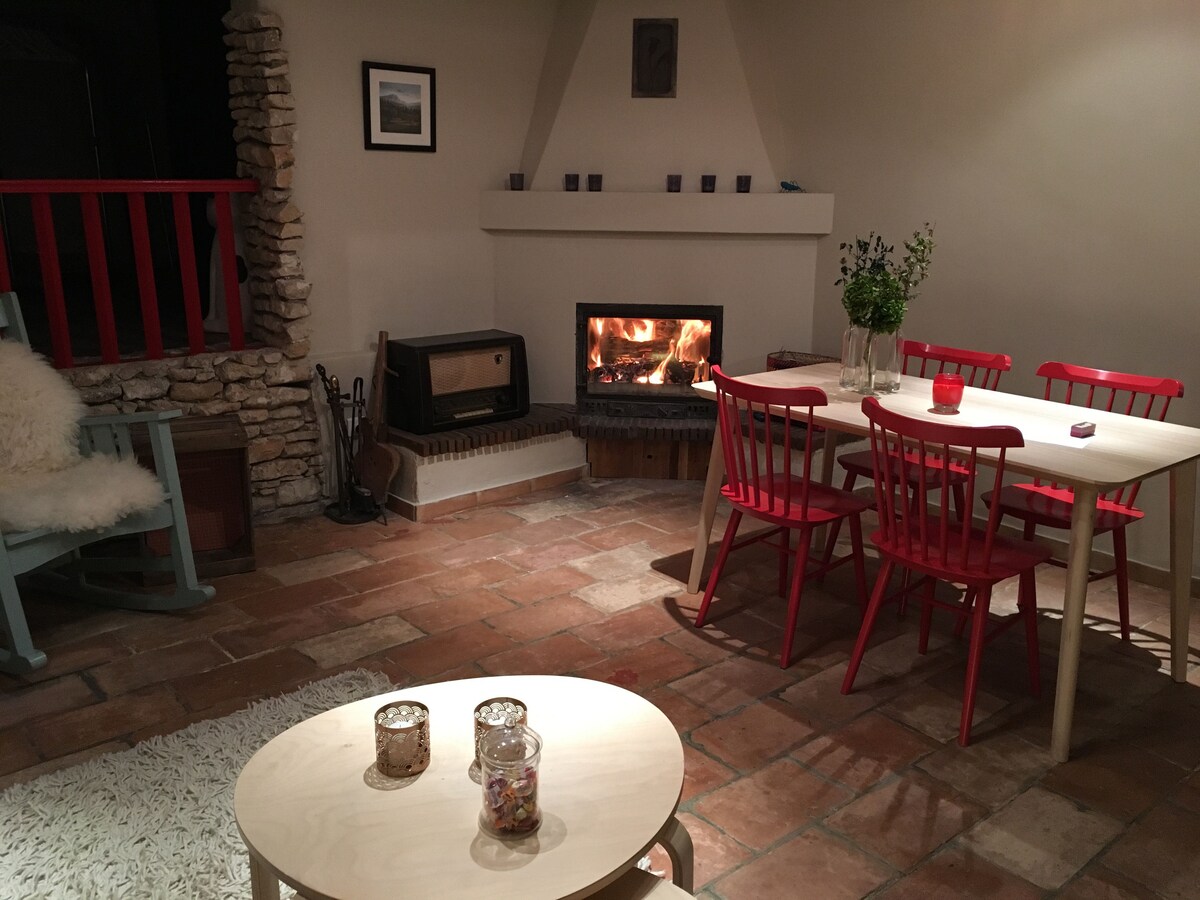 An inviting living area features a rustic stone fireplace with a warm fire. A wooden table surrounded by red chairs is positioned near the hearth. A light-colored rug lies under a round coffee table, creating a cozy space enhanced by soft lighting and simple decor.