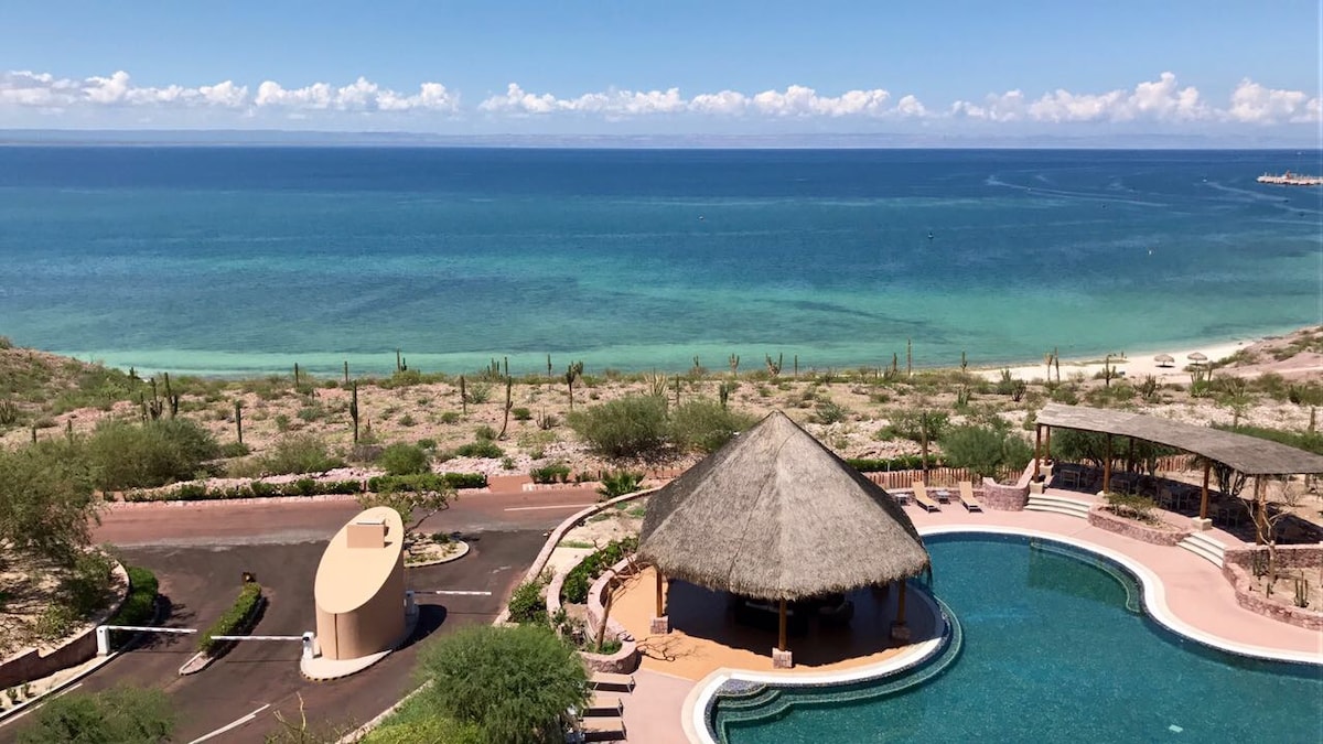 A spacious infinity pool is framed by tropical landscaping, leading to a pristine beach and azure ocean. A thatched-roof palapa provides shaded seating near the water, while gentle waves are visible in the distance. The scene is bathed in natural light beneath a clear sky.