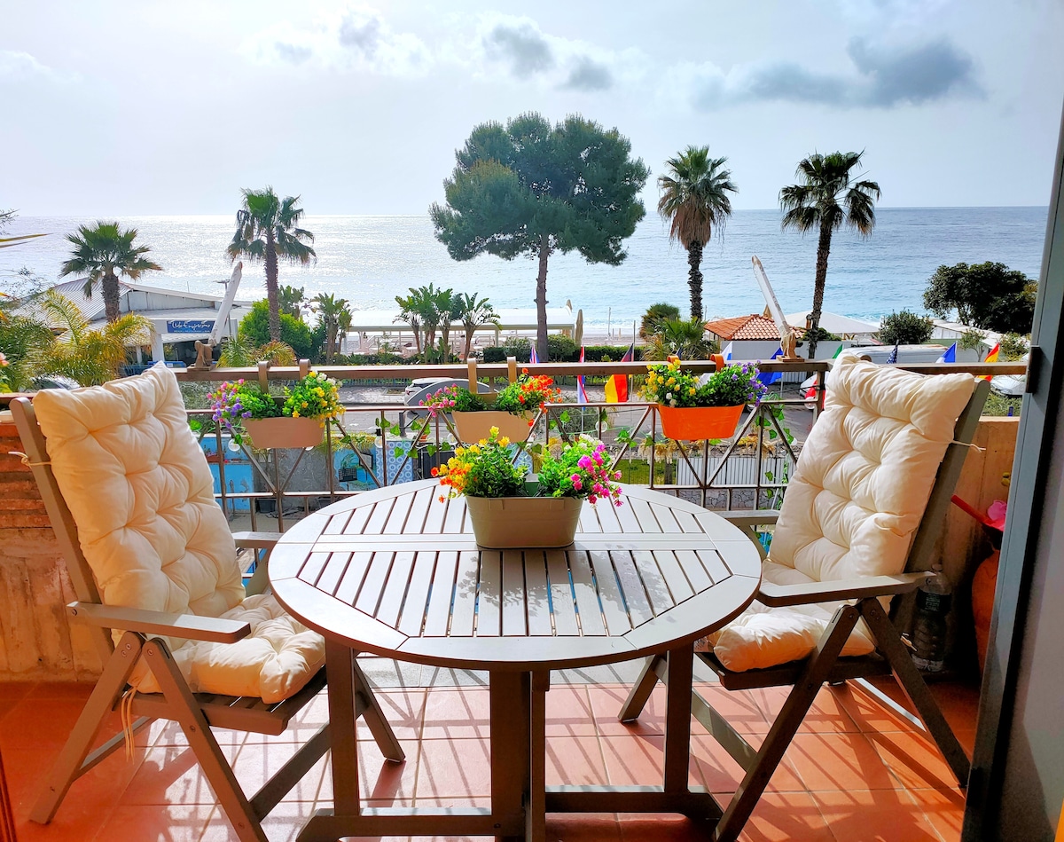A furnished terrace features a round table surrounded by two chairs, adorned with a planter overflowing with colorful flowers. The ocean and palm trees create a scenic backdrop, enhancing the serene atmosphere of outdoor relaxation.