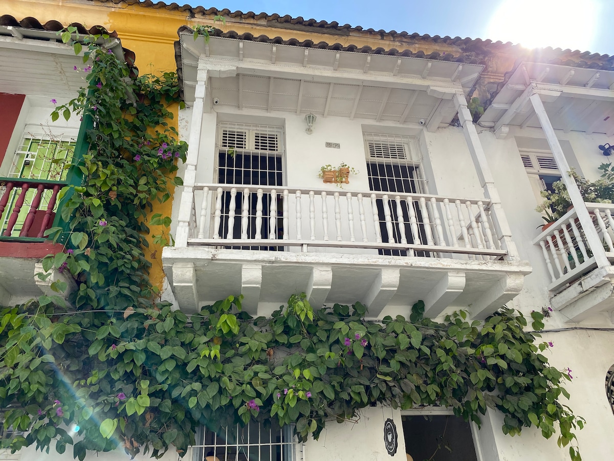 The duplex apartment features a charming balcony adorned with greenery. White railings encompass the covered area, while colorful walls painted in yellow and red bring vibrancy to the structure. Sunlight illuminates the scene, highlighting the lush vegetation that climbs along the building.