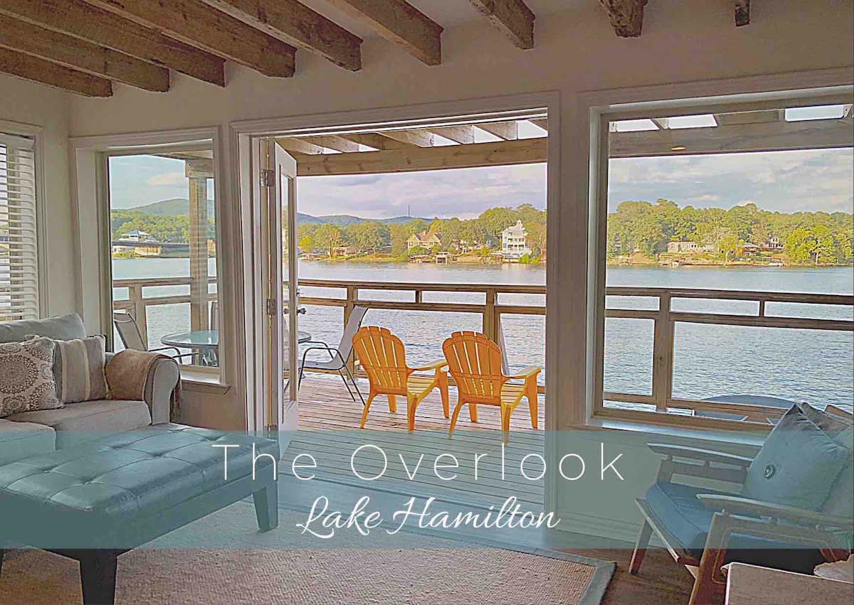 Large windows provide unobstructed views of Lake Hamilton, with light streaming into the living area. Two bright orange chairs sit on the balcony, inviting relaxation. A cozy seating area is visible inside, with soft furnishings and natural wooden beams accentuating the space.