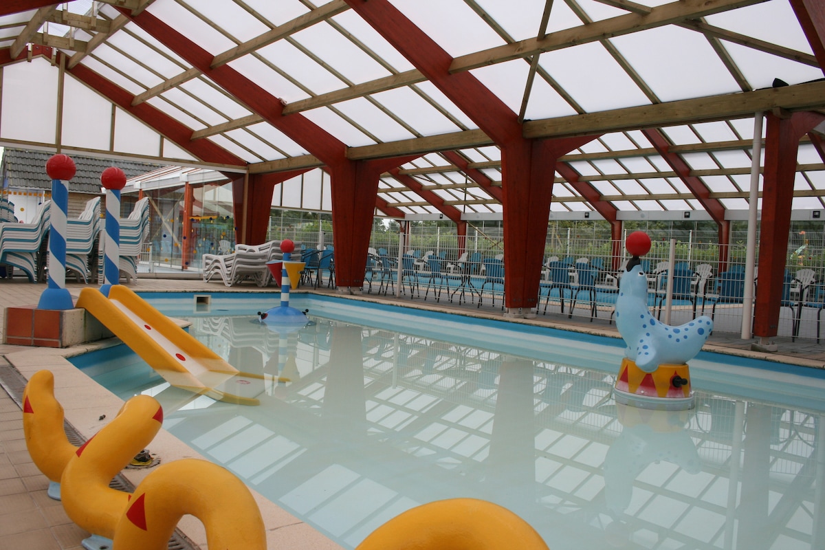 An indoor swimming pool features colorful slide structures and playful aquatic decorations. Sunlight filters through the transparent roof, illuminating the clear water. Surrounding the pool are rows of lounge chairs and tables, offering a relaxed space for visitors.