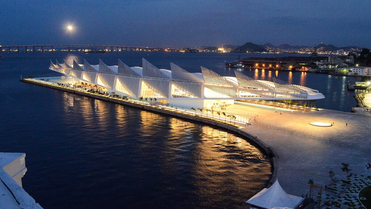 A modern architectural structure is illuminated against the night sky, reflecting on the calm waters below. The building features a unique design with numerous angular elements, while city lights twinkle in the distance, adding vibrancy to the scene.