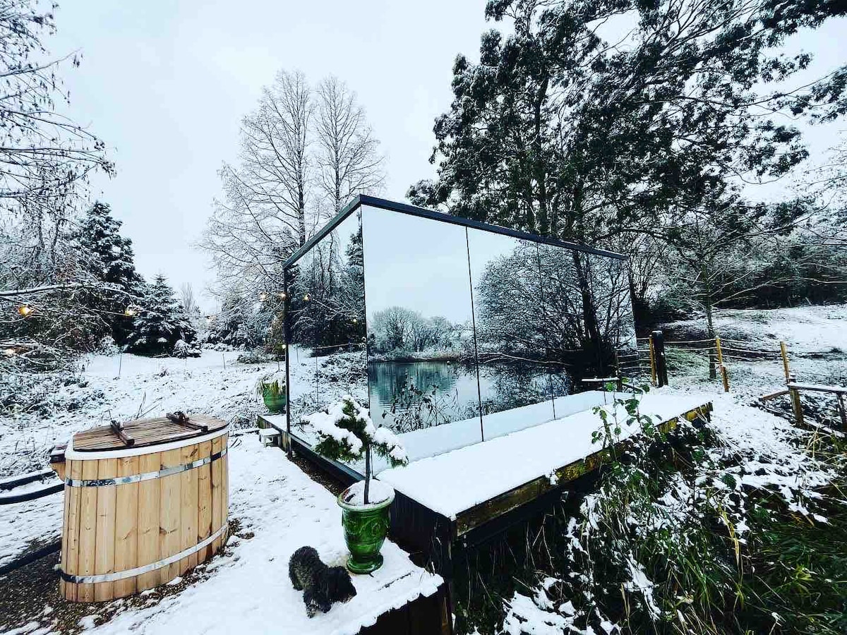 The ÖÖD House is set amidst a snow-covered landscape, featuring mirrored glass walls that reflect the surrounding winter scenery. A wooden hot tub stands nearby, and the serene lake can be seen through the glass, enhancing the peaceful atmosphere.