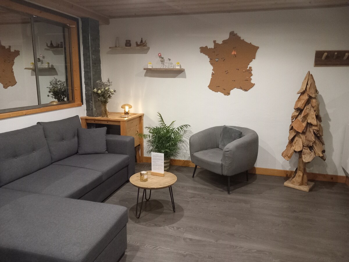 A cozy living area features a dark gray sectional sofa and a circular wooden coffee table. A small, round chair sits adjacent, while a floor lamp provides soft lighting. A decorative tree and wall art shaped like France add a touch of character to the space.