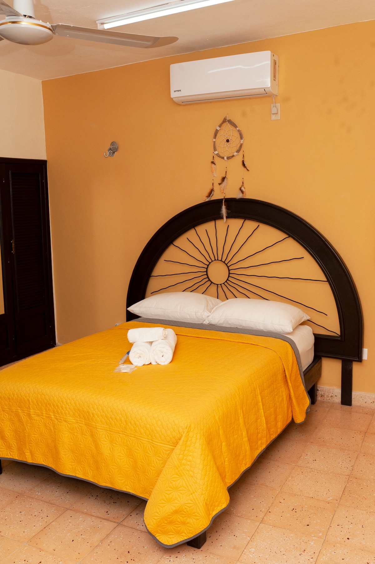 A bright sleeping area features a spacious bed adorned with a yellow quilt. Two neatly rolled towels are arranged on the bed. A wooden headboard with a sun design is positioned above the bed, complemented by a visible air conditioning unit on the wall.