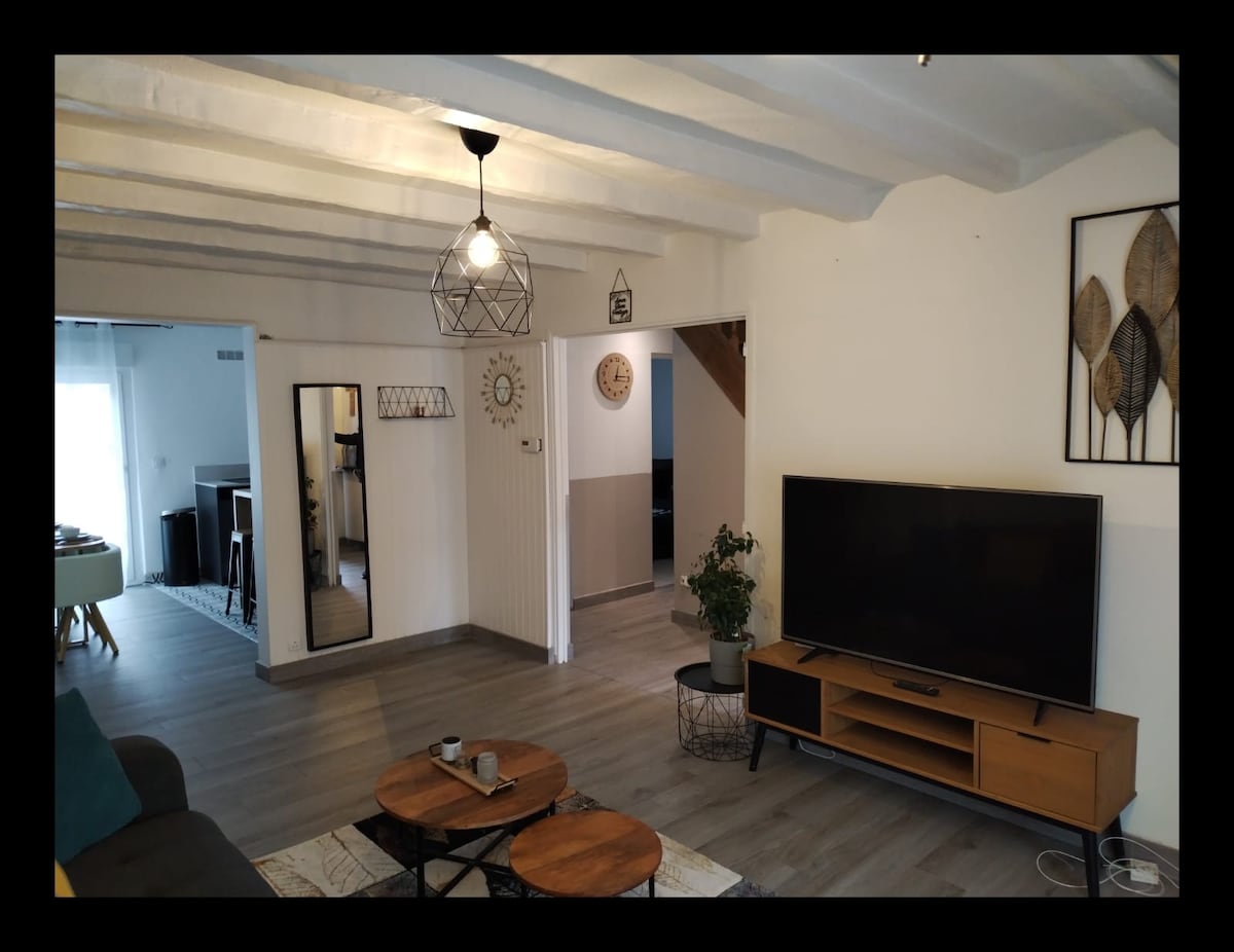 A spacious living area features modern wood flooring and exposed ceiling beams. A large television is positioned on a contemporary media console, complemented by two round coffee tables. A green potted plant adds a touch of nature, enhancing the overall ambiance.