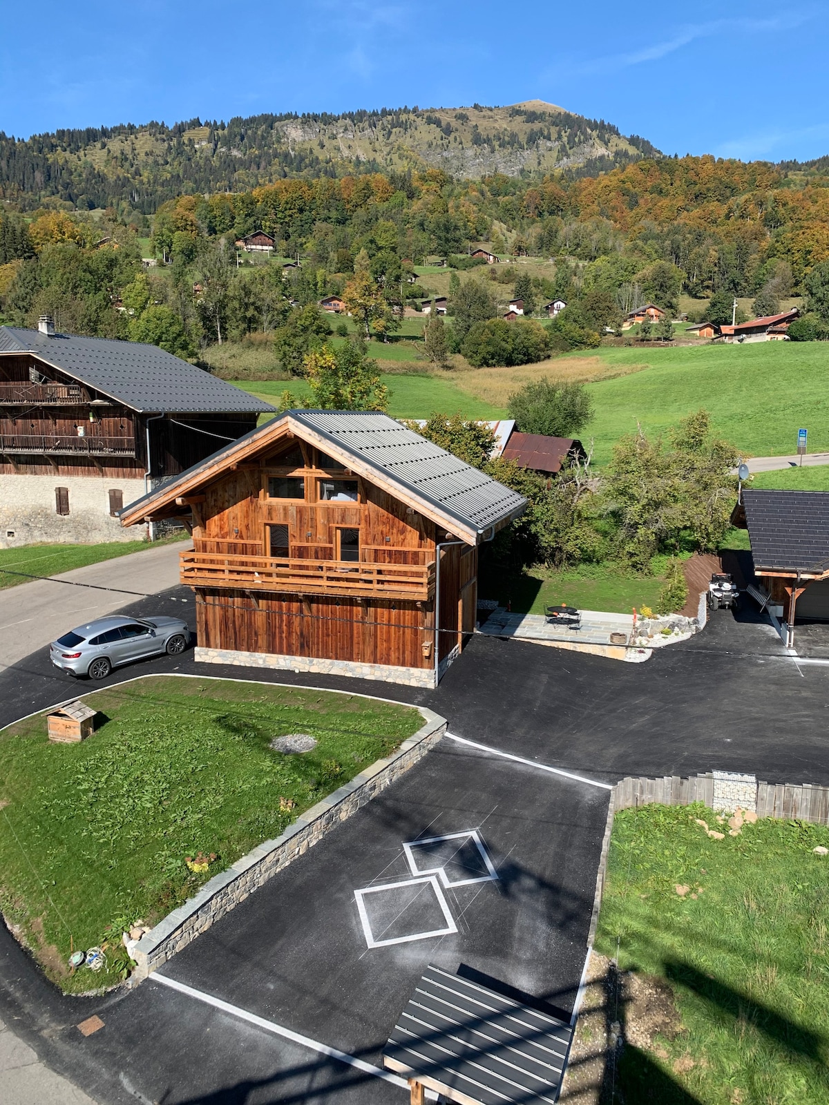 An aerial view of a rustic chalet features wooden exterior and a well-maintained garden. The parking area is clearly marked, accommodating vehicles. Surrounding lush greenery and distant mountains create a scenic backdrop, enhancing the tranquil environment of the property.