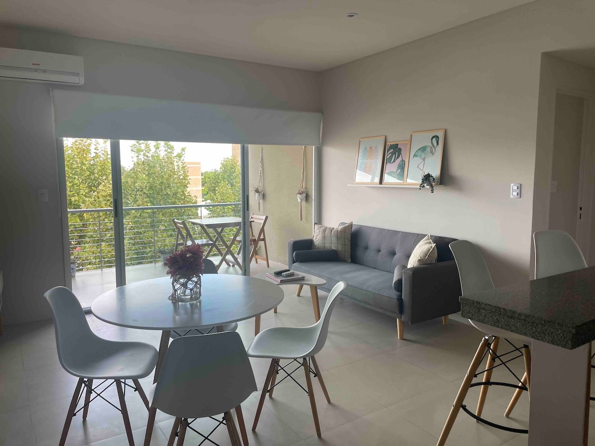 A modern living area is featured, showcasing a round dining table surrounded by white chairs and a comfortable gray sofa. Large windows allow natural light to fill the space, with greenery visible outside. Framed art pieces are displayed on the wall.