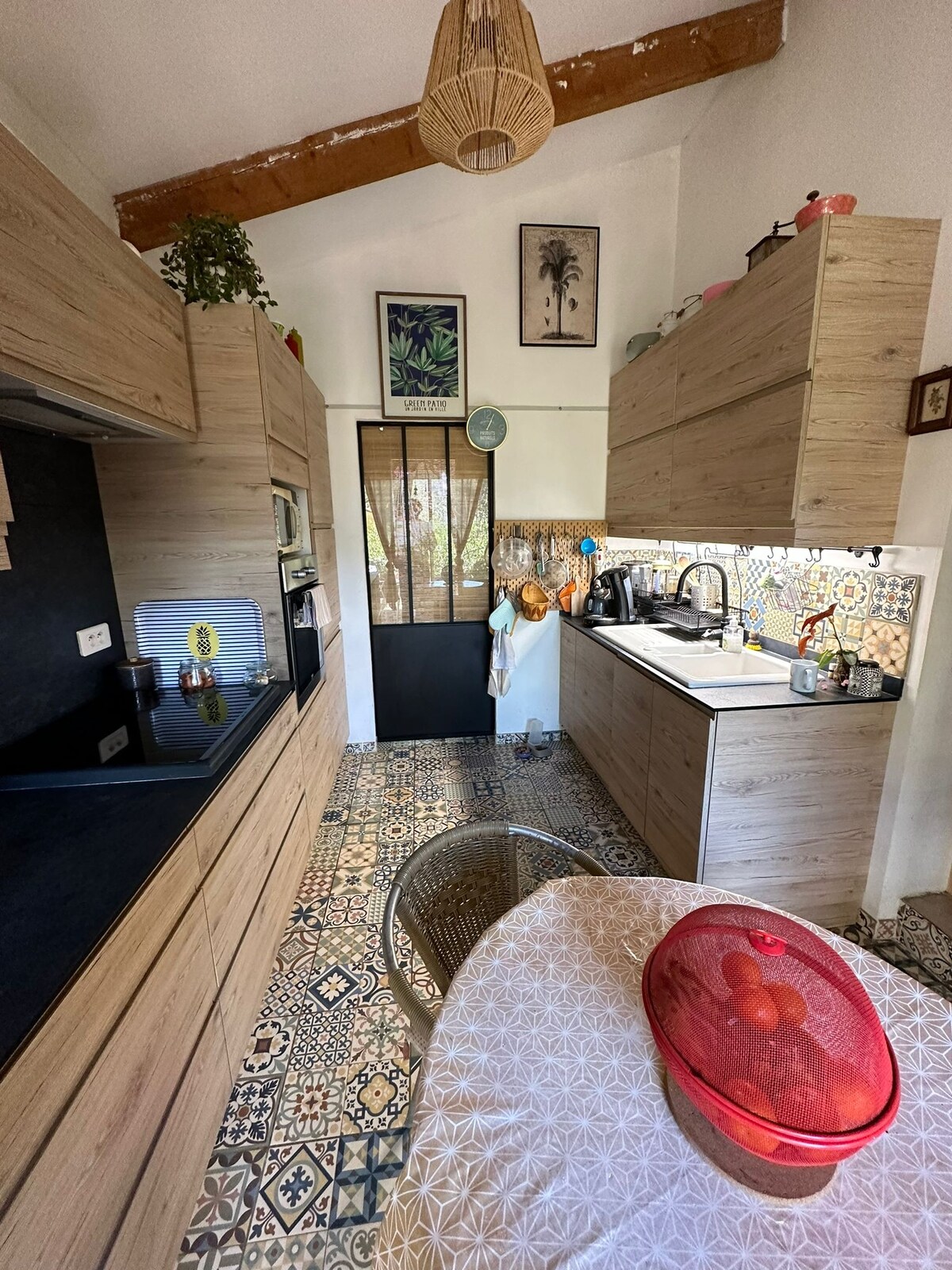 A well-equipped kitchen is showcased, featuring wooden cabinetry and a black countertop. Colorful tiles adorn the floor and backsplash, providing a vibrant backdrop. Natural light enters through a door leading to an outdoor area. A dining table is set with a bowl of fruit.
