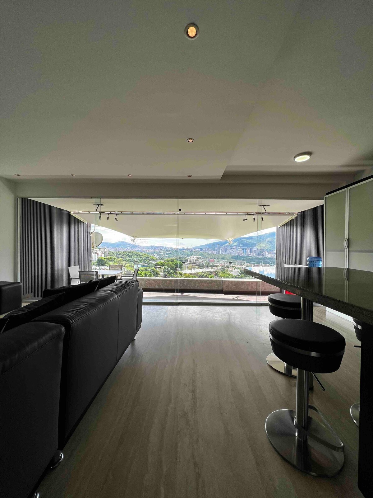 A modern living space is observed featuring a sleek kitchen bar and cozy seating area. Large glass doors open to a spacious terrace, offering panoramic views of the surrounding city and mountains. Natural light enhances the open layout, providing a welcoming ambiance.