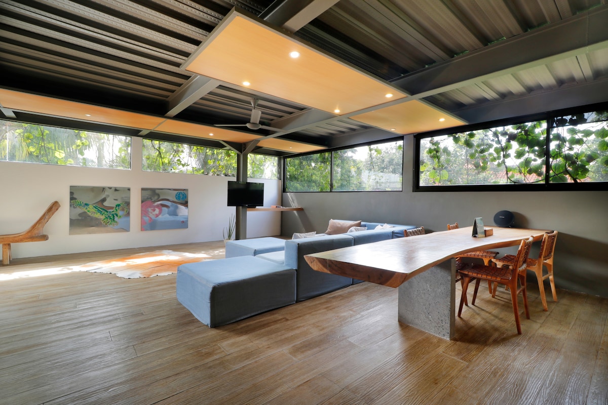 A spacious living area features large windows allowing natural light to fill the space. A massive wood dining table rests on a polished concrete leg. A comfortable sofa is arranged around the room, with stylish art displayed on the walls and a television visible in the corner.