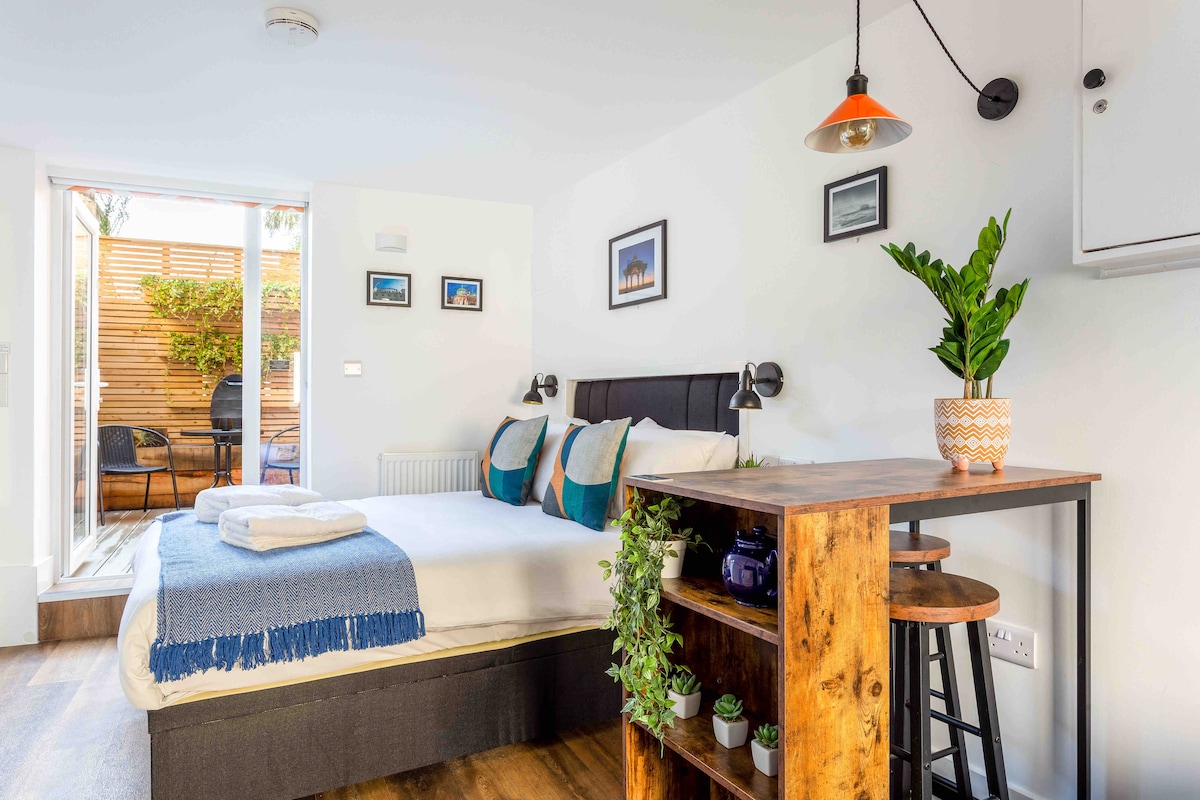 A spacious bedroom is highlighted by a stylish black bed with blue and green accent cushions. A wooden table with stools is set nearby, adorned with plants. Natural light enters through a door leading to a terraced garden, enhancing the relaxed ambiance.