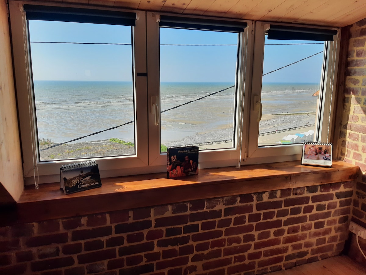 Three large windows offer a panoramic view of the sea, allowing natural light to fill the space. A wooden ledge displays photo calendars, while the exposed brick wall adds a touch of texture to the room.