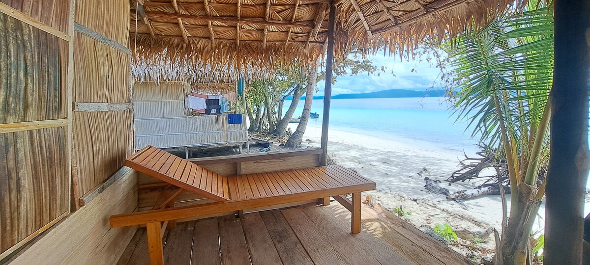 A shaded wooden lounging area features a simple, slatted sunbed facing the beach. The surrounding natural elements include palm trees and a sandy shore, creating a serene coastal environment. Gentle waves are visible in the background, enhancing the tranquil setting.
