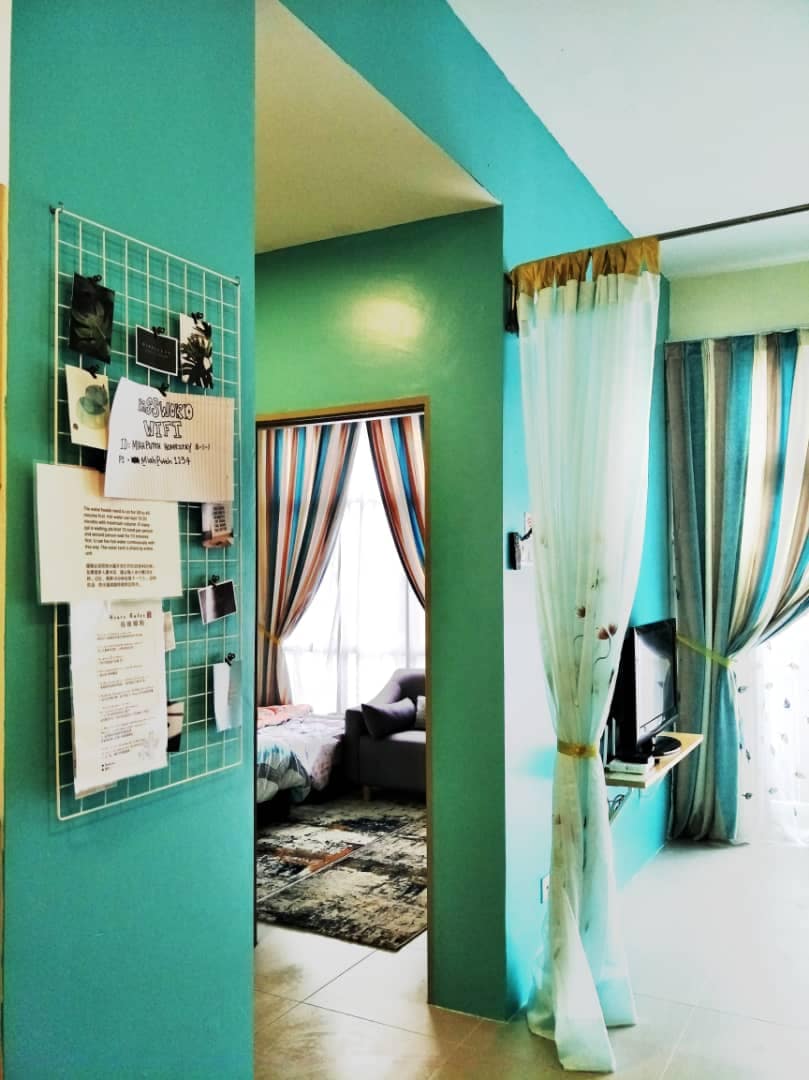 A turquoise hallway leads into the living area, adorned with colorful curtains framing a large window. A television is mounted on a wall opposite a vibrant area rug. A bulletin board hangs on the wall, displaying various notes and information.