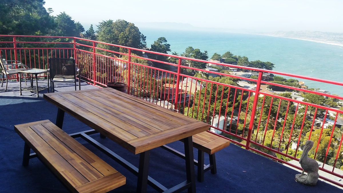 An outdoor dining area features a wooden table and benches set against a backdrop of sweeping ocean views. The railing provides safety while allowing for unobstructed sightlines of the coastline and hills. Greenery surrounds the space, creating a natural ambiance.