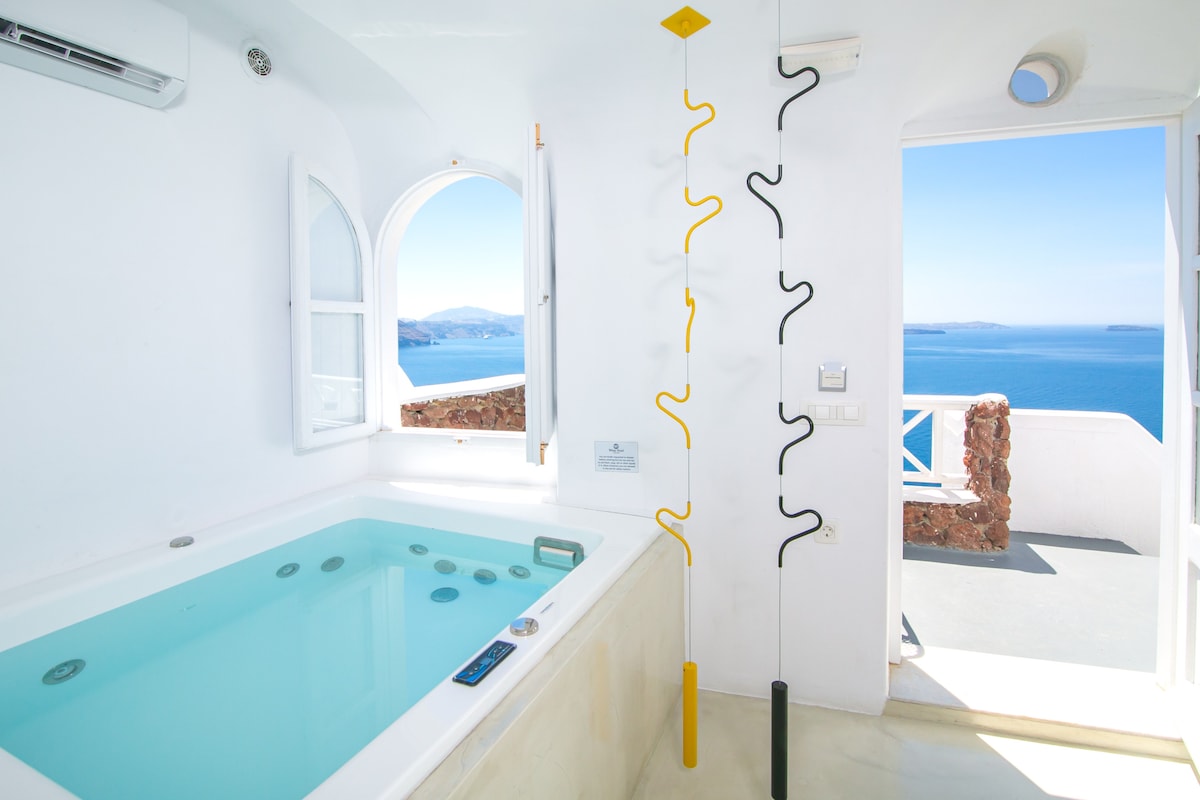 An indoor jacuzzi is positioned by an arched window, offering views of the Aegean Sea. The space features white walls and light-colored flooring, with sleek fixtures attached to the wall providing a contemporary design element.