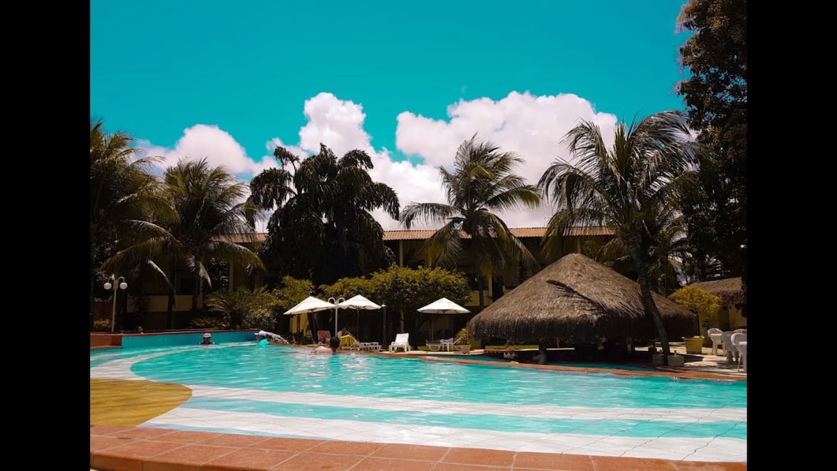 A large swimming pool is surrounded by swaying palm trees, sun loungers, and shaded umbrellas. A thatched-roof structure provides a tropical vibe. Bright blue skies with fluffy white clouds add to the outdoor serenity, creating a relaxing atmosphere by the water.