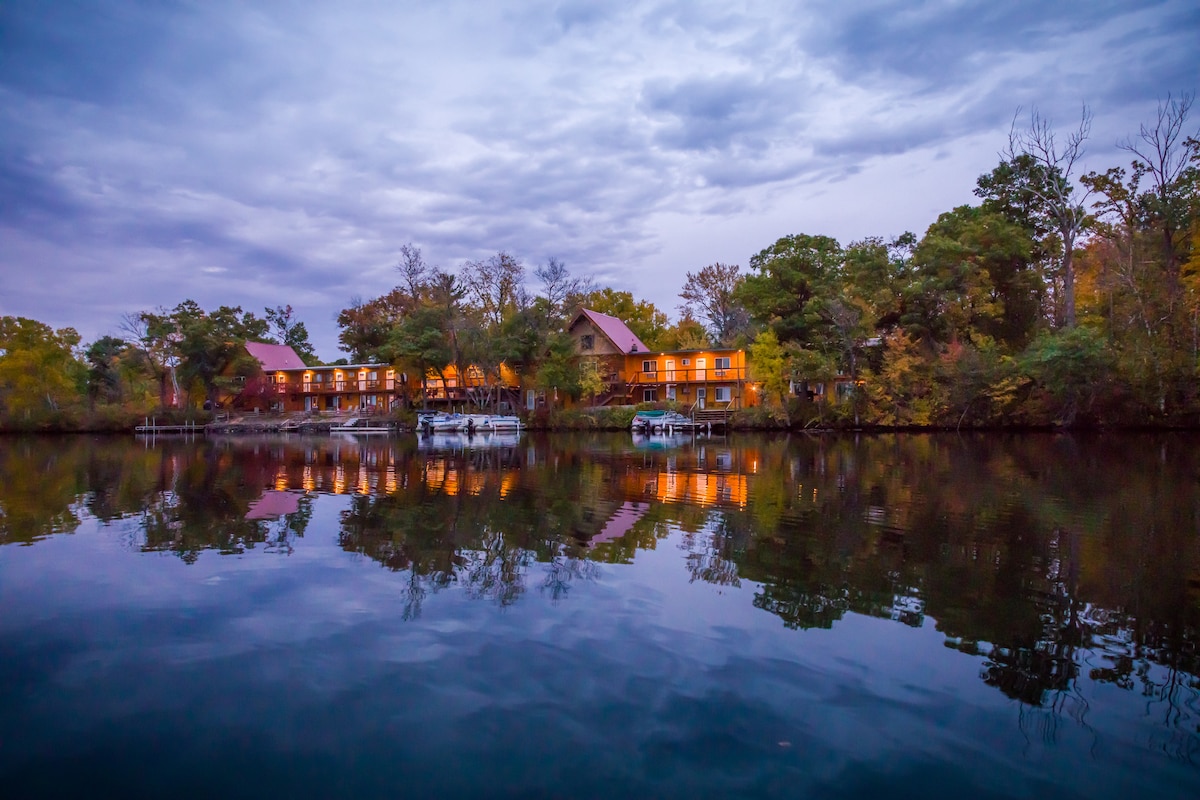 The tranquil lakeside scene features an orange-hued building reflecting on calm waters, surrounded by greenery. The sky displays soft clouds at dusk, adding to the serene atmosphere. Docks and boats are visible along the water's edge, enhancing the natural setting.
