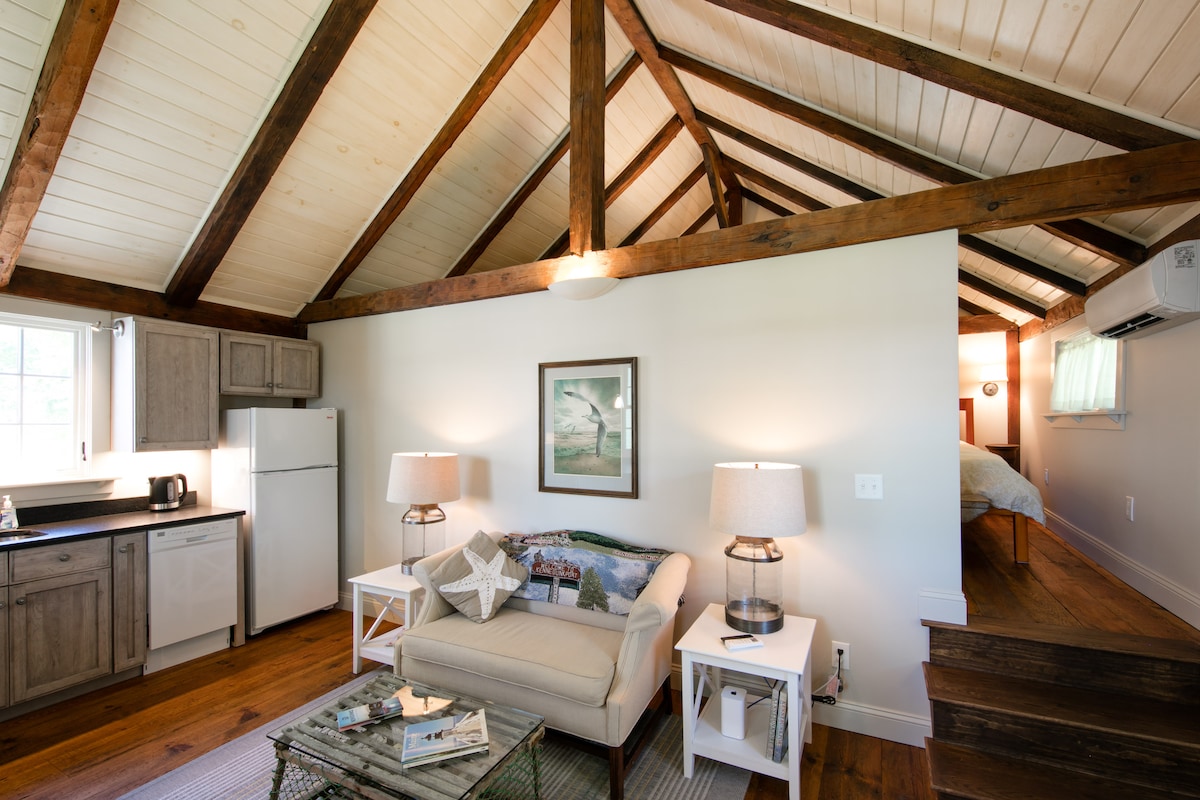The open concept living area is illuminated by natural light streaming through the windows, complemented by wood-beamed ceilings. A cozy sofa is arranged with decorative pillows, while a coffee table displays a selection of books. A small kitchenette is positioned nearby, featuring a refrigerator and sink.