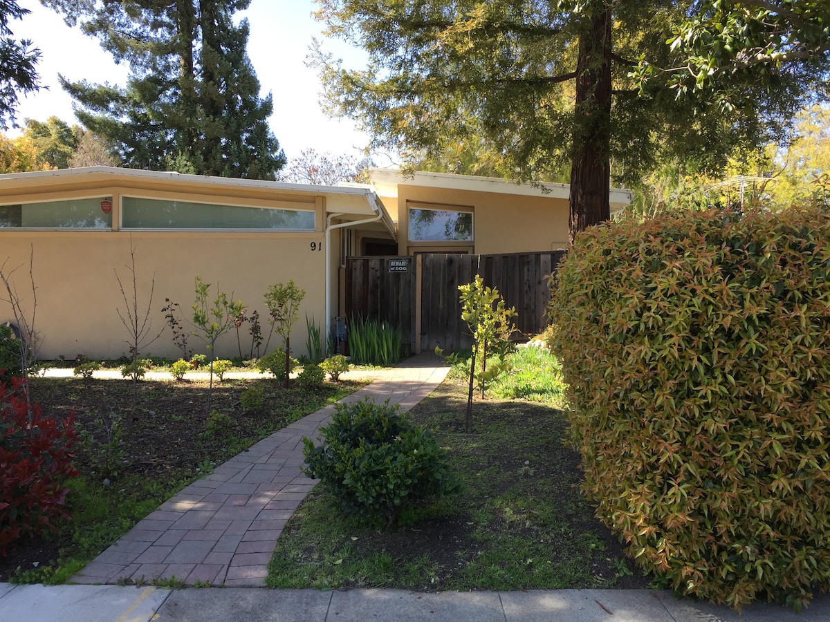 The image displays the exterior of a well-maintained property set amidst lush greenery. A path of square paving stones leads to a gated entrance, flanked by neatly trimmed bushes and vibrant foliage. Large windows hint at a spacious interior behind the beige stucco facade.
