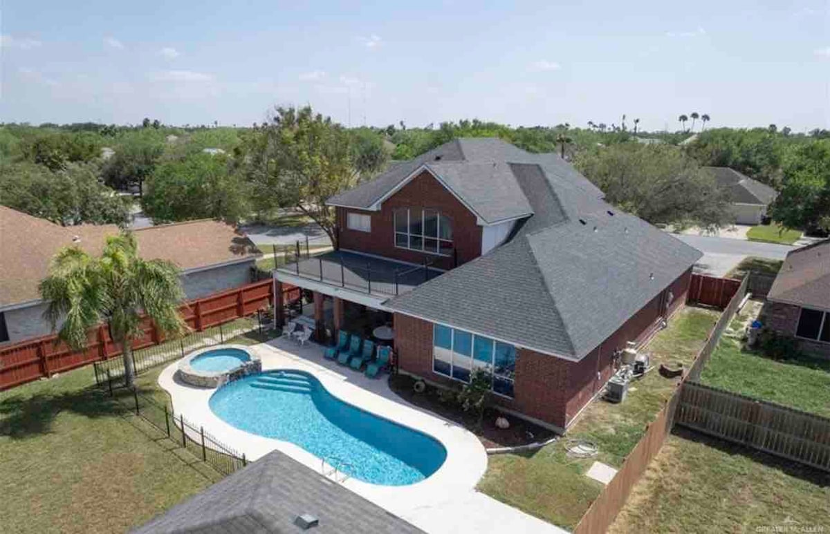 An aerial view of a spacious backyard reveals a large swimming pool, surrounded by a stone deck and seating areas. A shaded patio is located adjacent to the pool, and the property features a vibrant green landscape with trees and fencing enclosing the area.