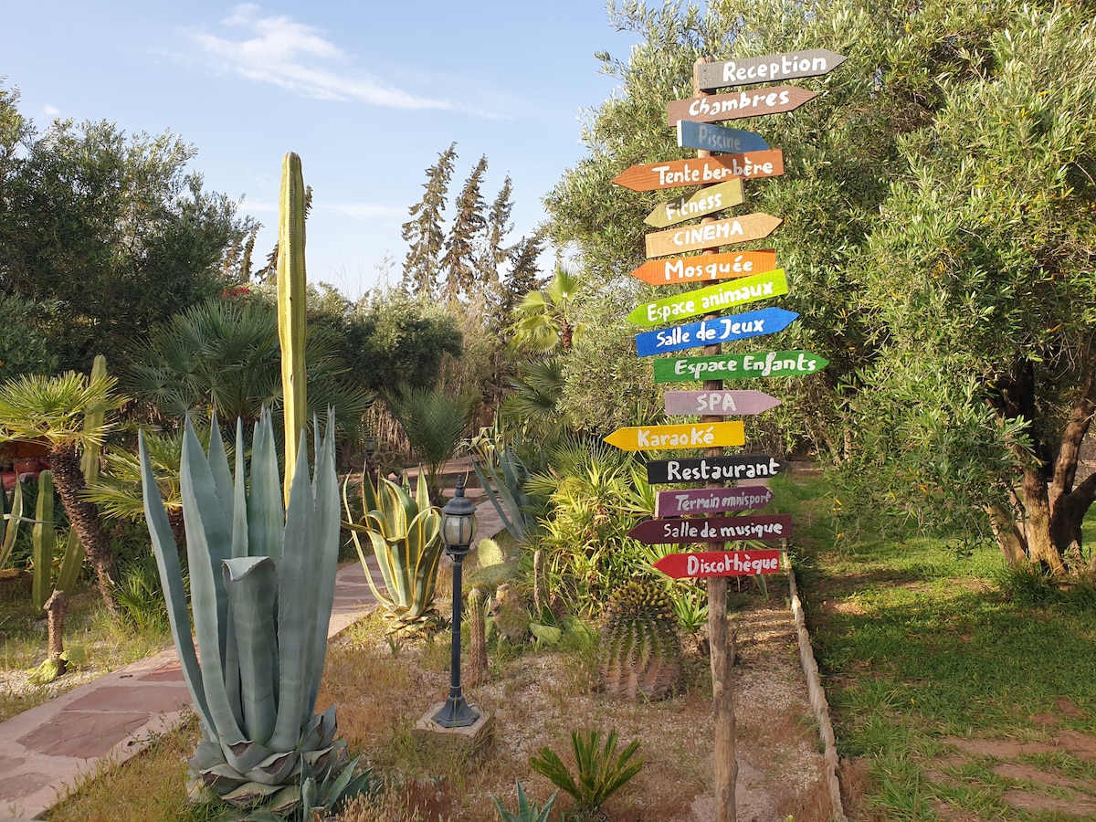 A vibrant pathway is lined with directional signs, indicating various amenities including the reception, restaurant, and spa. Lush greenery surrounds the signs, with cacti and other plants creating a natural backdrop, enhancing the inviting atmosphere of the outdoor space.