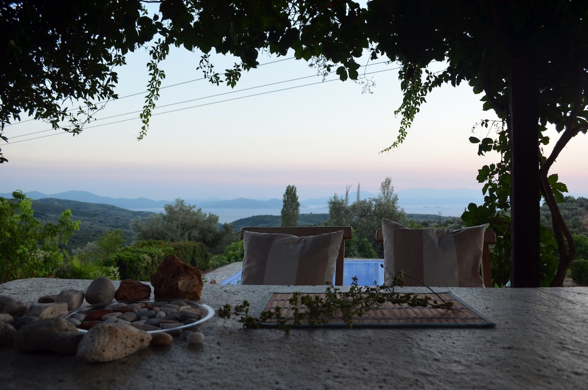A tranquil outdoor view is presented, featuring a stone table adorned with small pebbles and plants. In the background, two chairs are positioned under a canopy of greenery, offering an expansive vista of rolling hills and the distant sea at dusk.
