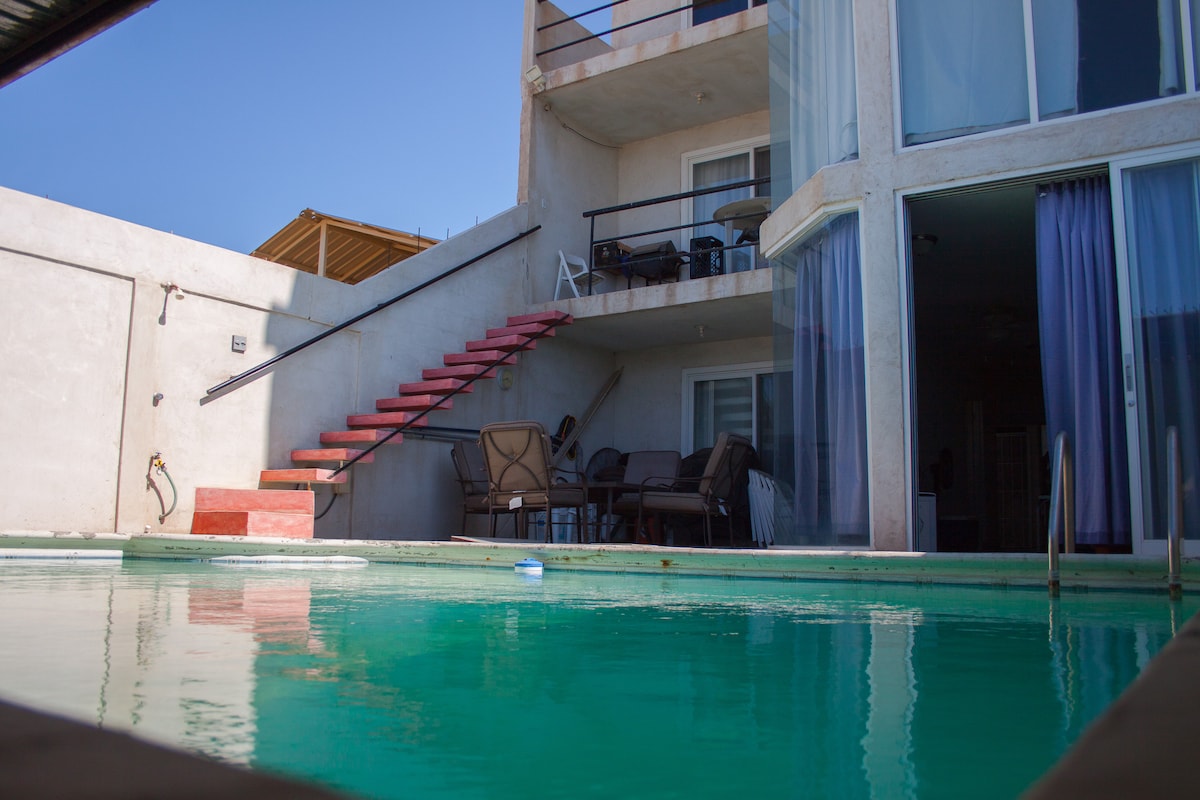 A private swimming pool reflects sunlight, with steps leading to an outdoor seating area and a two-story condominium in the background. Large glass doors provide a view into the living space, while vibrant blue curtains add a touch of color.
