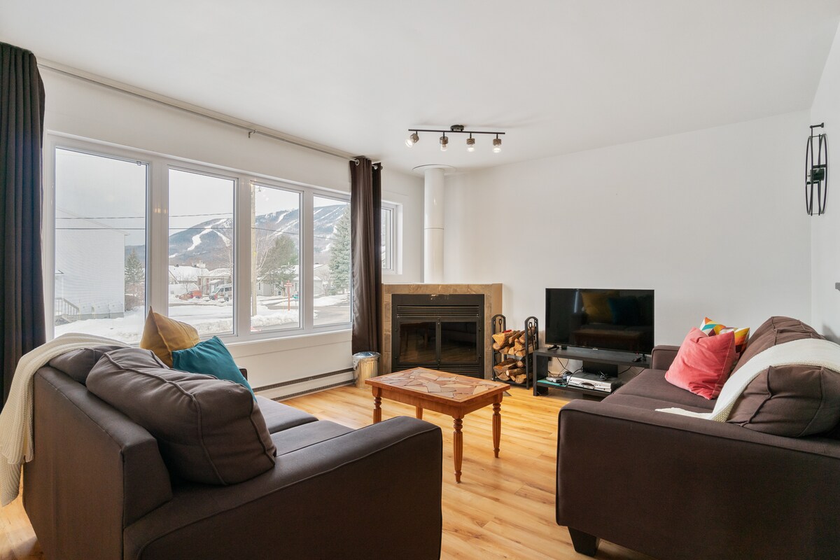 A bright living area features large windows that provide views of snow-covered mountains. Comfortable sofas surround a wooden coffee table, and a television is positioned near the corner. A fireplace adds a focal point to the space, enhancing the cozy atmosphere.