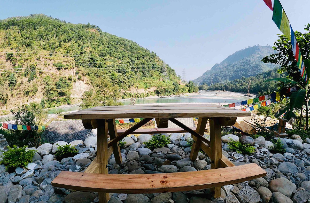 A wooden picnic table is positioned on a rocky surface, overlooking a river bordered by lush greenery and hills. Colorful prayer flags gently flutter in the breeze, enhancing the serene natural setting. Sunlight illuminates the scene, creating a peaceful atmosphere.