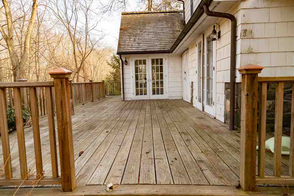 A spacious wooden deck is presented, surrounded by wooden railings. French doors lead into the cottage. Natural light fills the area, enhanced by the surrounding trees. The surface of the deck shows a natural wood finish, indicating a well-maintained outdoor space.