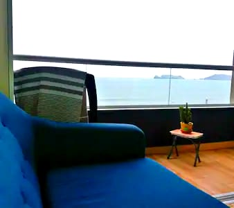 A blue sofa is positioned in a cozy living area, facing large windows that provide a view of the ocean. A small wooden table with a potted plant is placed beside the sofa, enhancing the tranquil setting.