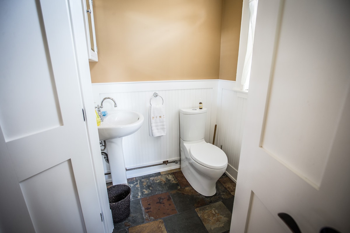 A bright and clean bathroom is shown, featuring a white sink and a modern toilet. The walls are painted a warm beige, complemented by white wainscoting. Natural light filters in through a window, illuminating the slate flooring.