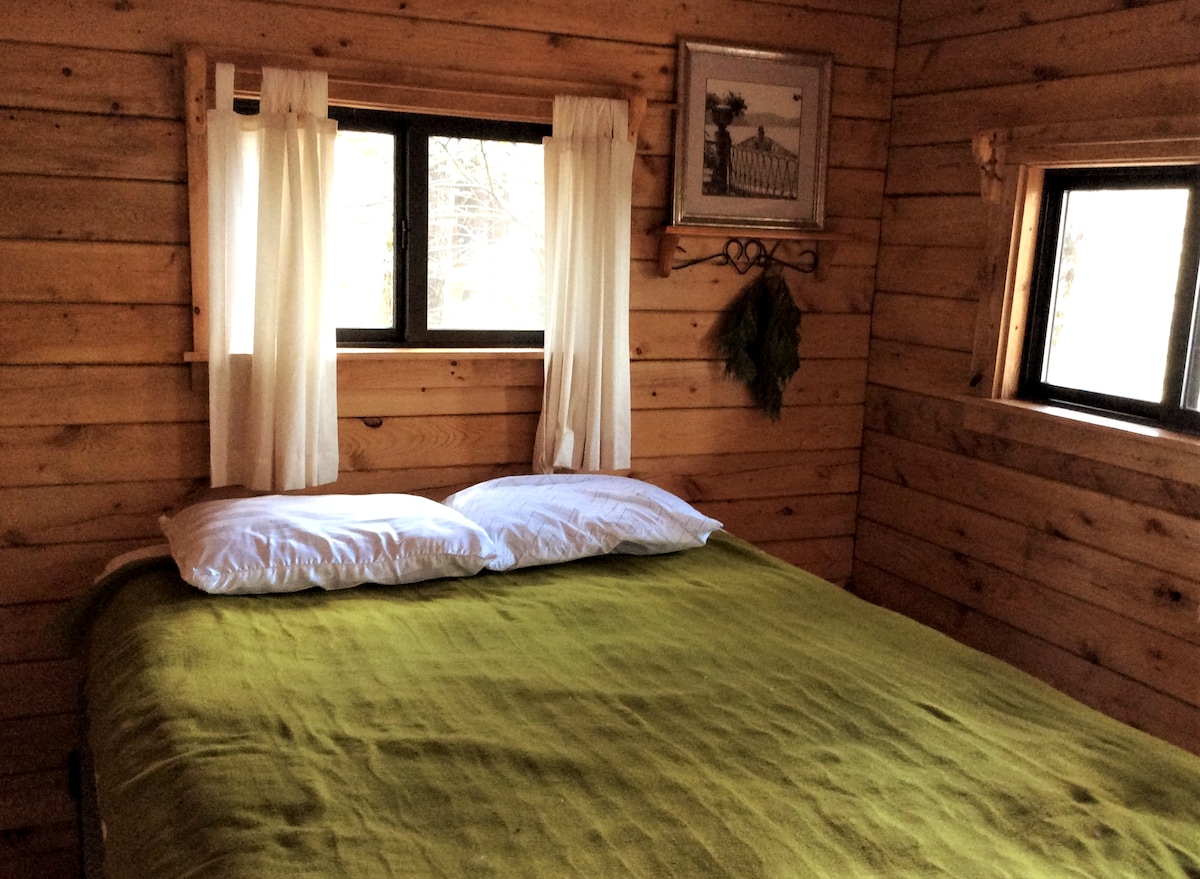 A cozy sleeping area features a spacious bed with a green blanket and two white pillows. Two windows allow natural light to brighten the wood-paneled walls, enhanced by light-colored curtains. A simple piece of wall art is displayed above the bed.