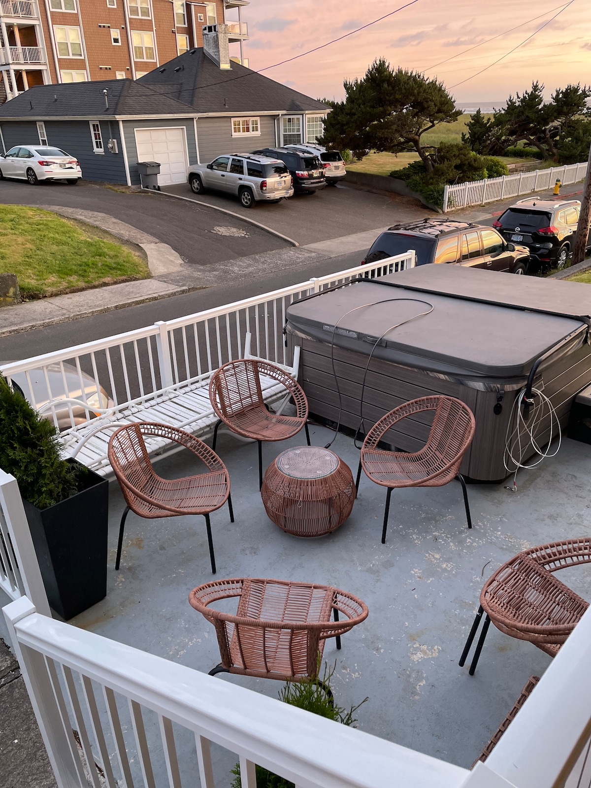 An outdoor seating area features a hot tub and six woven chairs surrounding a round table. The surface is paved, and greenery is visible in planters. A sunset casts warm light, enhancing the view of the neighborhood and nearby homes.