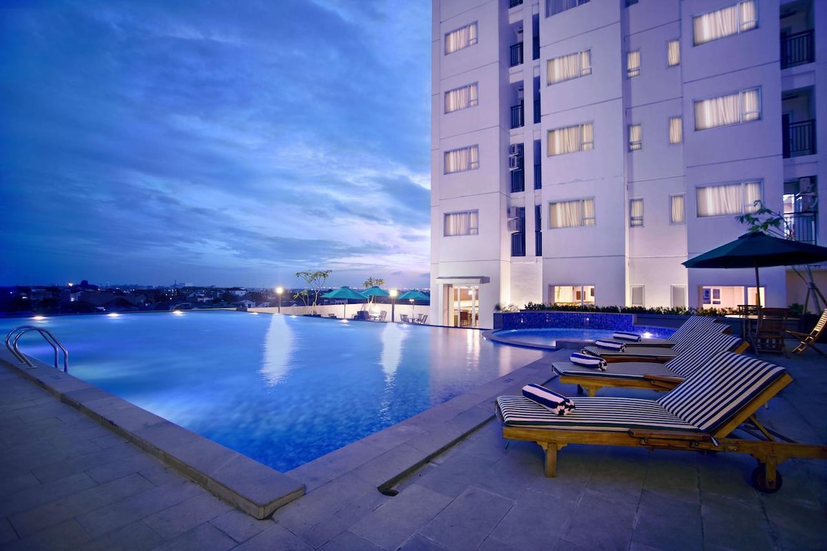 An inviting pool area is framed by the apartment building, featuring several lounge chairs arranged along the edge. The water reflects the evening sky, and an umbrella to shade guests is visible beside the pool. Soft lighting from surrounding windows enhances the tranquil ambiance.