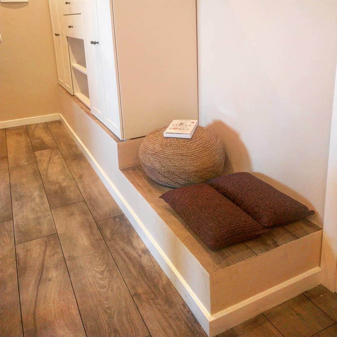 A cozy seating area features a soft, textured pouf alongside two plush cushions on a low wooden platform. Light-colored walls and natural wood flooring create a serene environment. A small book rests open on the platform, inviting relaxation.