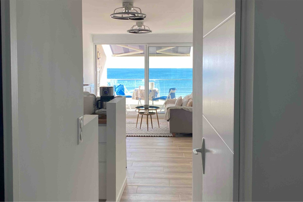 A bright entryway leads into the living space, framed by large sliding glass doors that reveal stunning ocean views. Light floods the room, highlighting the modern furnishings and a cozy seating area. A dining table sits adjacent, creating a welcoming atmosphere.