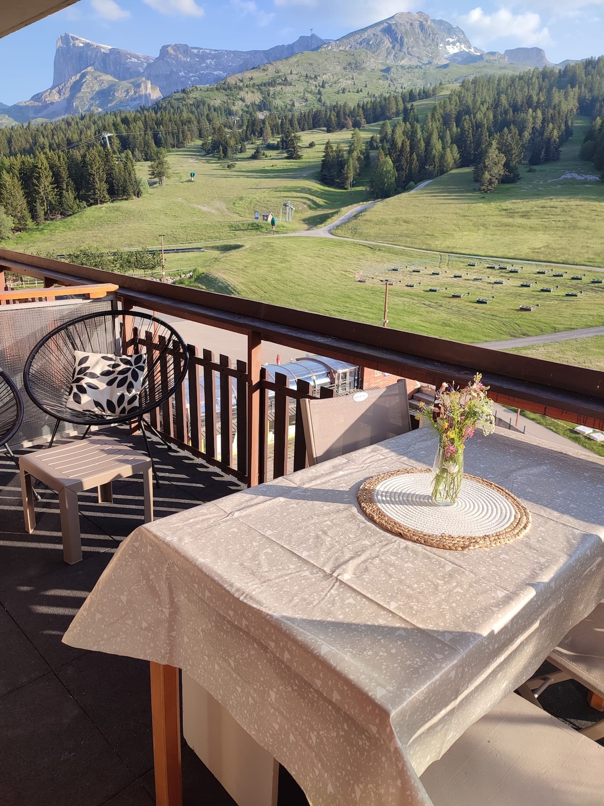 A balcony features a table set with a beige tablecloth, accompanied by a chair and a decorative vase holding fresh flowers. The surrounding landscape showcases green mountains, trees, and ski slopes under a clear sky, creating a serene outdoor space.