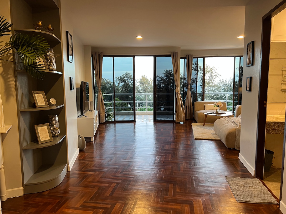 A spacious living area features polished wooden flooring and large glass windows that offer expansive views of the surrounding landscape. A modern sofa is positioned alongside a minimalist television stand, enhancing the room's openness and inviting natural light.