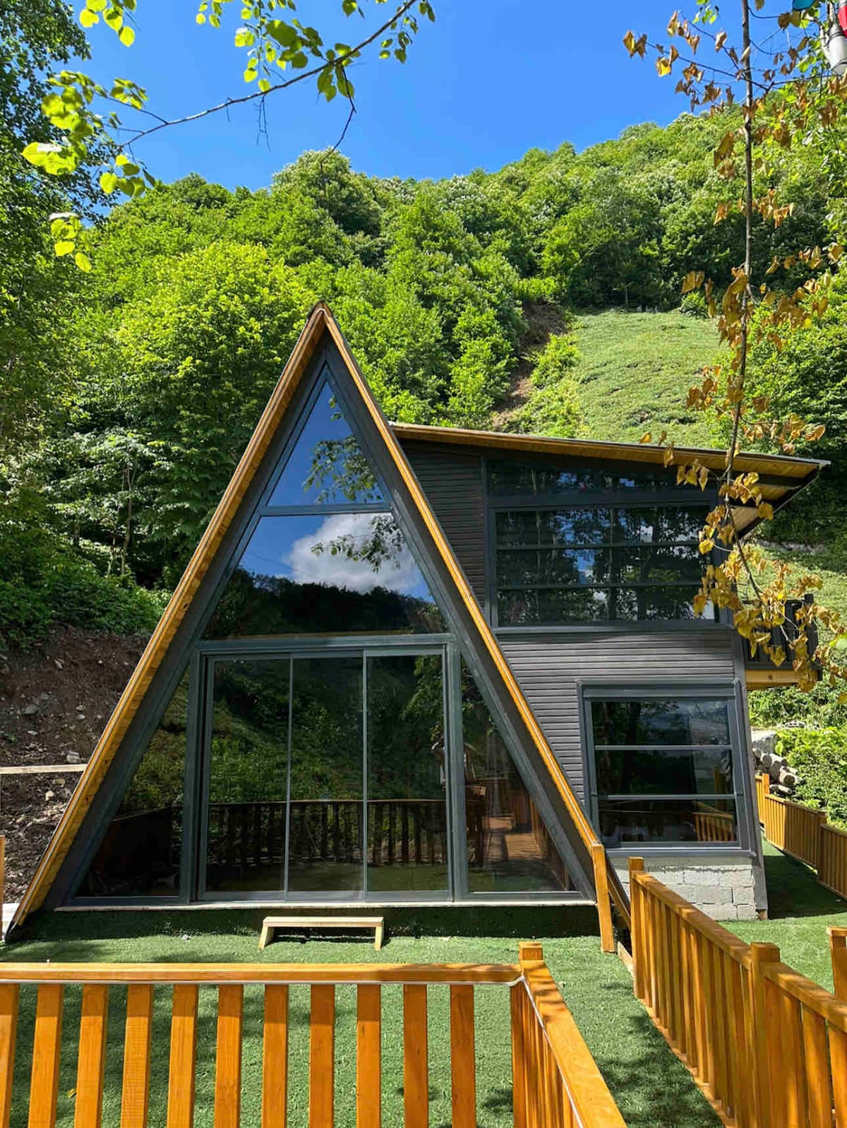 An A-frame structure is nestled in a lush green environment, featuring large glass windows that provide ample natural light. A wooden deck extends outward, framed by a low wooden fence. Surrounding trees and hillside create a serene backdrop.