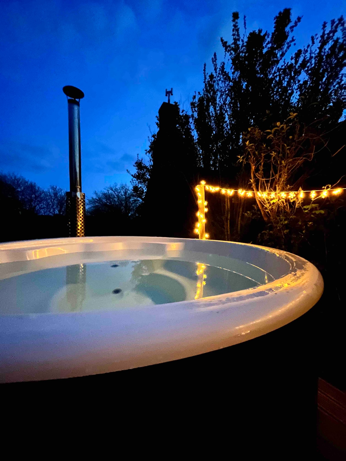 A wood-fired hot tub is positioned in a peaceful garden setting, surrounded by lush greenery. Soft, warm lights illuminate the area, enhancing the tranquil evening ambiance. The still water reflects the twilight sky, creating a serene atmosphere for relaxation.