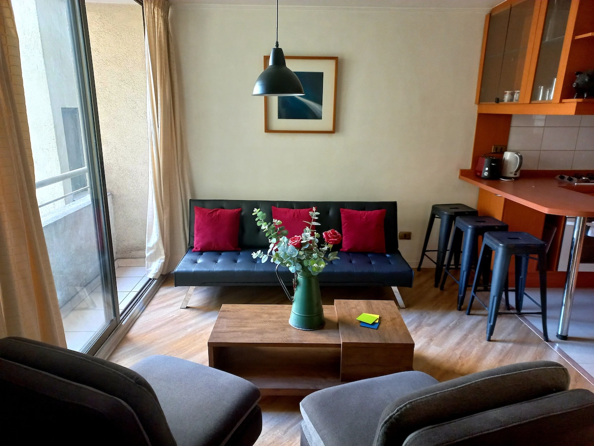 A living area is presented with a dark futon adorned with red cushions, paired with a wooden coffee table. A vase of flowers provides a focal point, while the kitchen area features bar stools and appliances. Natural light filters through the glass balcony door.
