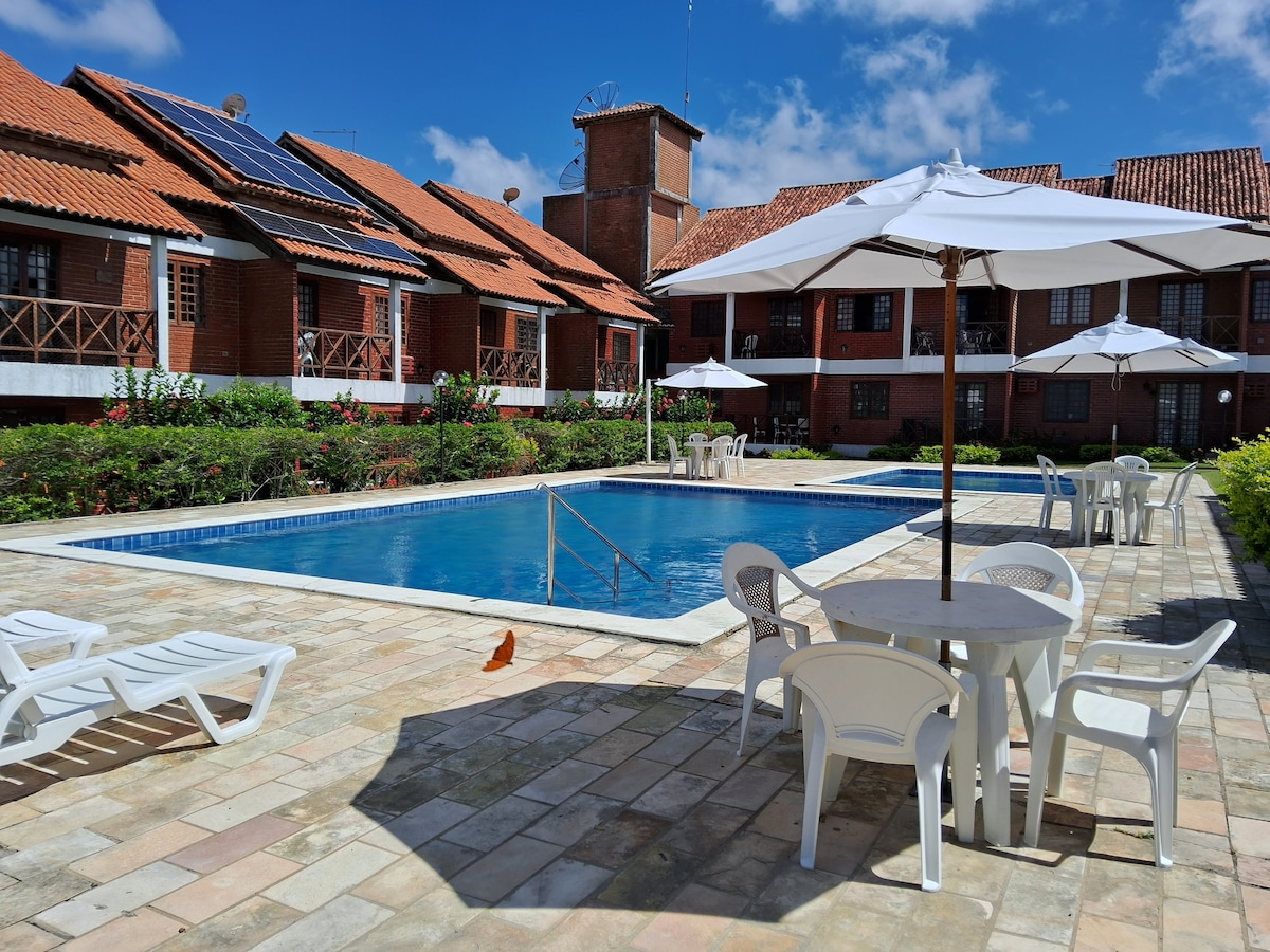 An outdoor pool area is surrounded by neatly arranged lounge chairs and tables under white umbrellas. The pool is framed by lush greenery and the brick buildings exhibit a traditional architectural style. The sky is clear, enhancing the tranquil atmosphere.