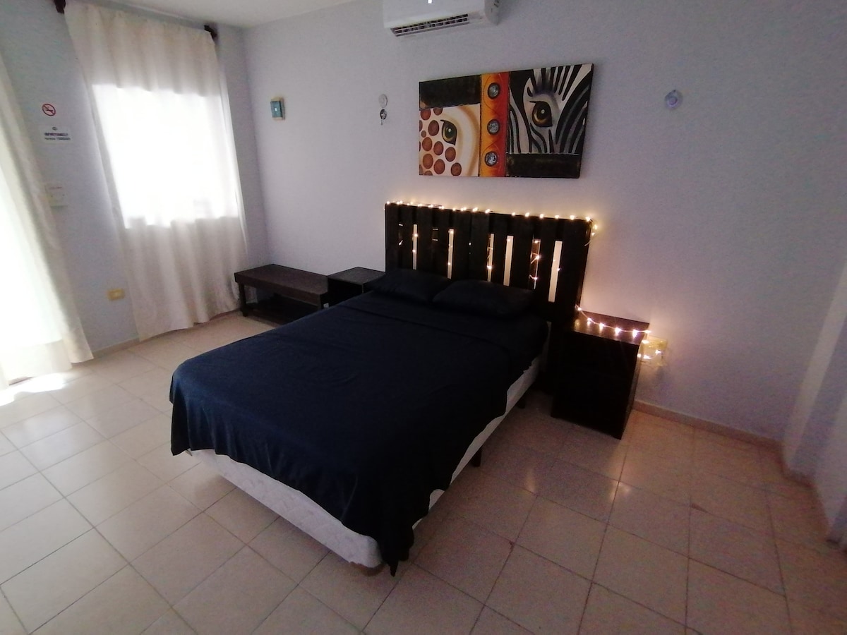A spacious bedroom features a dark bed frame with a double bed, accompanied by a small nightstand. A soft glow from string lights adds warmth to the space. A large window drapes sheer curtains, allowing natural light to fill the room. Simple wall art enhances the calm ambiance.